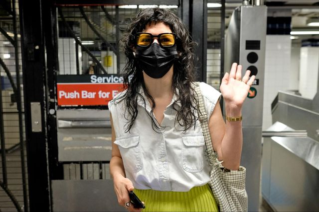A woman stands in front of turnstiles. She is wearing a mask and is waving.